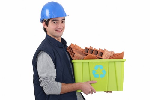 A young man wearing a blue safety helmet and a navy work jacket with grey sleeves is holding a light green recycling bin with a blue recycling symbol on the front. Inside the bin, there are neatly stacked red clay bricks with rough, textured surfaces and rectangular shapes, some with small holes through the centre. The man is standing against a plain white background, smiling slightly, and facing slightly to the right. The scene suggests a professional waste collection or rubbish removal service, with a focus on building materials or construction debris. This visual relates to commercial waste removal, potentially for a client in Millbank or nearby areas, highlighting the handling of materials suitable for disposal or recycling by companies like Commercial Waste Millbank.