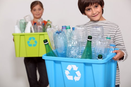 A young boy with dark hair and a striped shirt is holding a blue recycling bin filled with clear plastic bottles of various sizes, some with caps, arranged upright and leaning against each other. Behind him, a young girl with light brown hair is holding a green recycling container filled with similar plastic bottles, positioned partially out of focus. The scene takes place indoors against a plain light-colored background, emphasizing the focus on waste recycling. The plastic bottles in both containers are transparent, with different textures and shapes, indicating a collection for recycling purposes. The bright colors of the bins, blue and green, contrast with the neutral tones of the children’s clothing and backdrop, subtly aligning with waste management activities common in Millbank and broader London areas. Commercial Waste Millbank's services are supported by this visual emphasis on plastic bottle recycling, reflecting environmentally mindful rubbish collection. The lighting is even and neutral, highlighting the containers and their contents clearly and professionally.