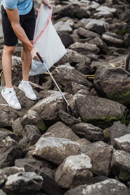 A shoreline strewn with various types of litter, including plastic bottles, crumpled plastic bags, paper debris, and small fragments of waste, all resting on a mixture of rocks, soil, and plant materi