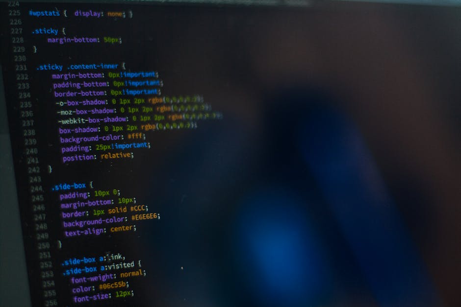 A person's hand, wearing a wristwatch with a patterned strap, is positioned on the keyboard of a silver laptop resting on a wooden surface. The laptop screen displays lines of code or text in a dark b