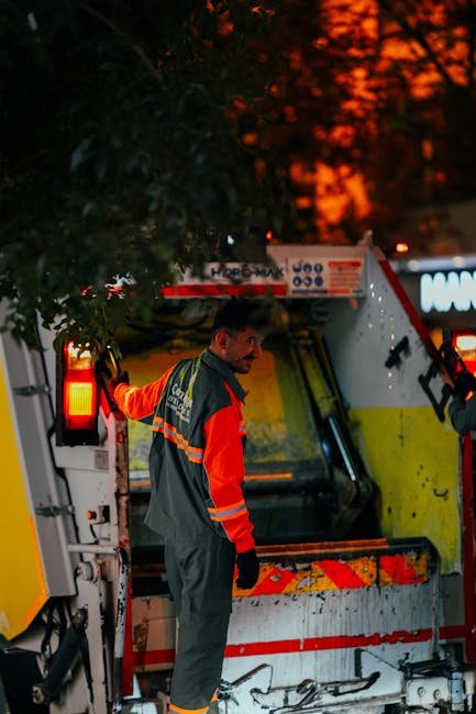 A waste collection worker is standing next to a large street-level refuse collection vehicle during early evening or nighttime, with a background of orange-red sunset sky visible through the trees. Th