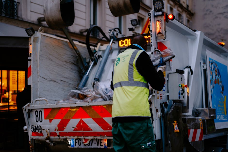 A waste collection worker is standing next to a large street-level refuse collection vehicle during early evening or nighttime, with a background of orange-red sunset sky visible through the trees. Th